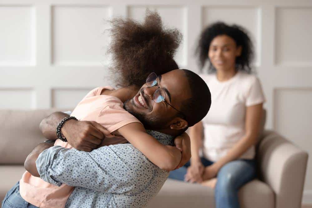 Image of a happy African American dad embracing his daughter while cuddling at home, featuring The Jimenez Law Firm and highlighting 'What Are the Forms of Joint or Shared Custody in Texas?', ensuring it aligns with the page's context.